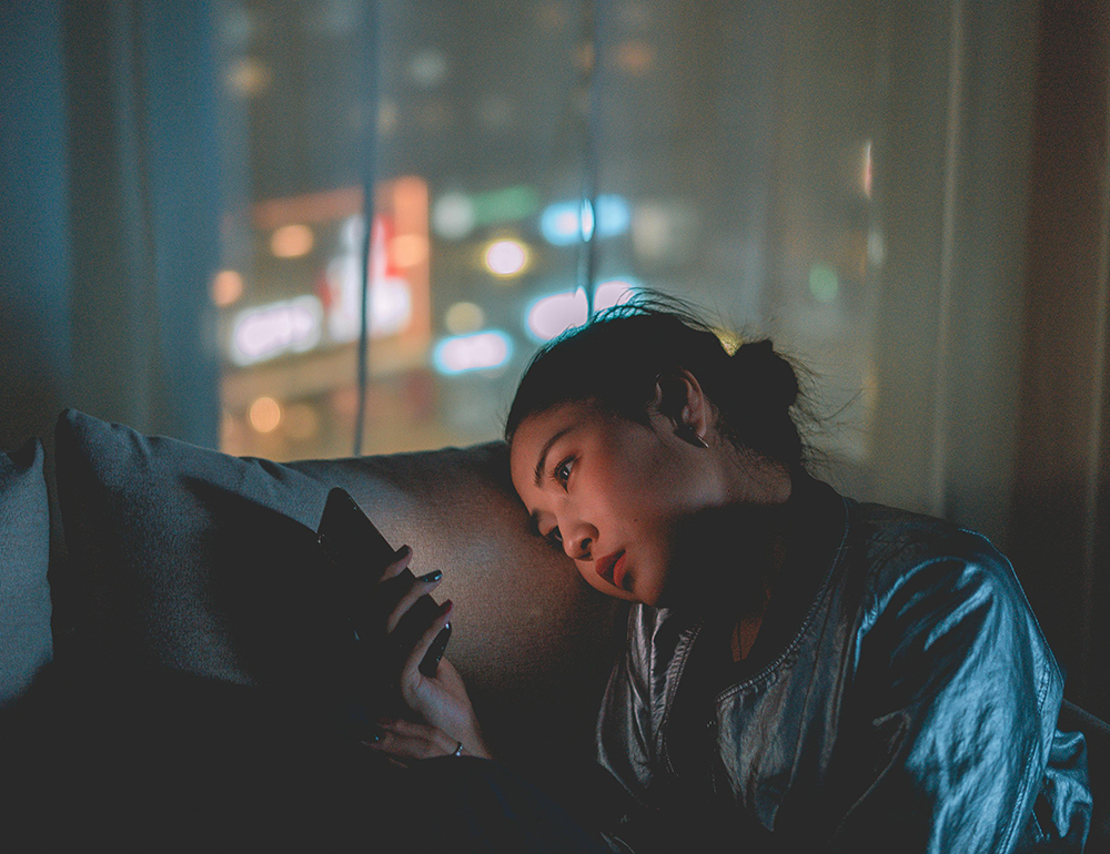 A young woman stares at her phone while sitting in a dimly lit room, capturing the emotional toll of an anxious avoidant relationship.