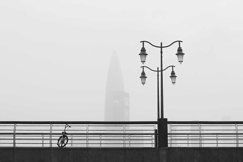 A foggy cityscape featuring a distant skyscraper, a lone bicycle leaning on a bridge railing, and twin street lamps – an image evoking the psychology of ghosting.