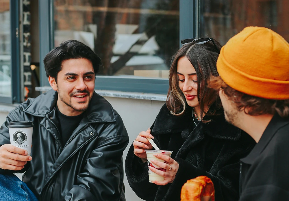 Three friends sit together outside a café, chatting warmly while enjoying coffee and pastries.