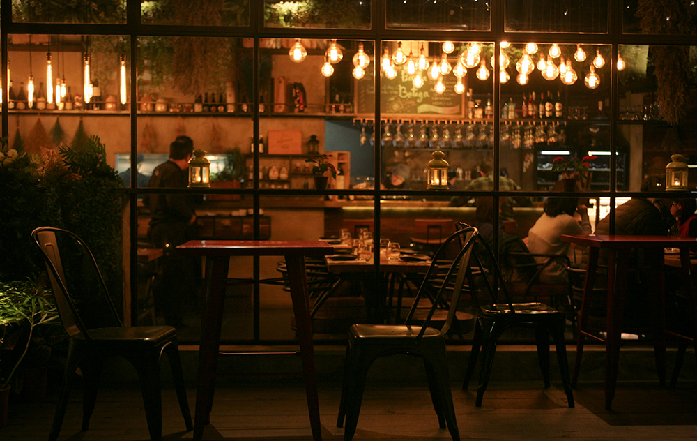 Warmly lit restaurant interior viewed through large windows, with empty chairs outside and diners enjoying conversations inside.