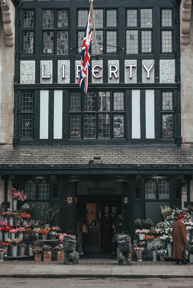 A street-level view of the historic Liberty department store in London, featuring a Union Jack flag, floral displays and Tudor-style architecture.