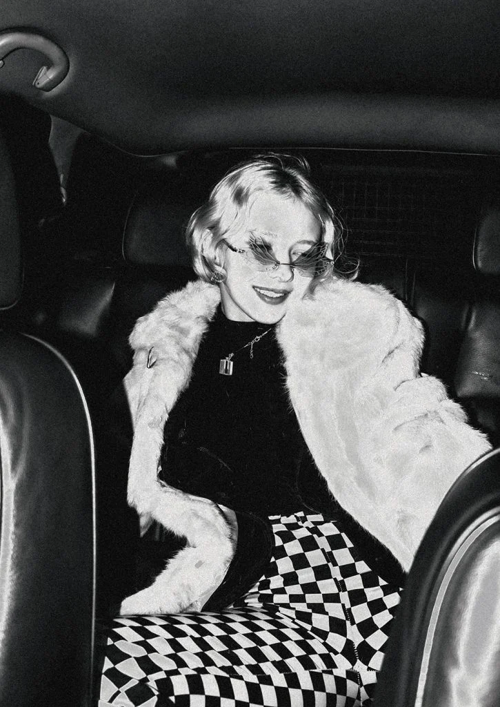 A woman with short blonde hair wearing sunglasses, a faux fur coat and chequered trousers sits smiling in the backseat of a car at night.