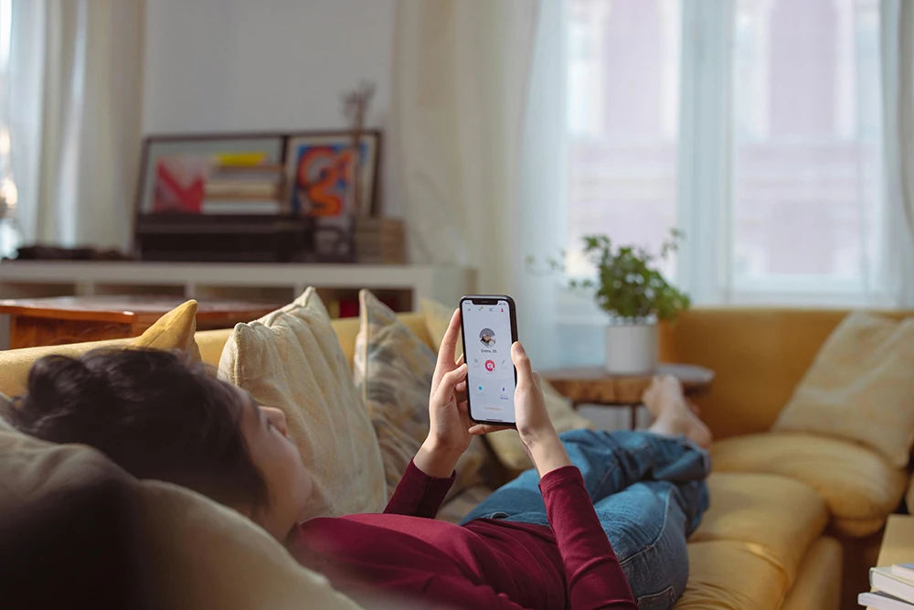 A person lying on a sofa holds a smartphone, scrolling through a dating app profile.