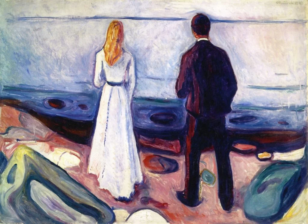 A man and woman stand side by side on a rocky shore, looking out at the sea.