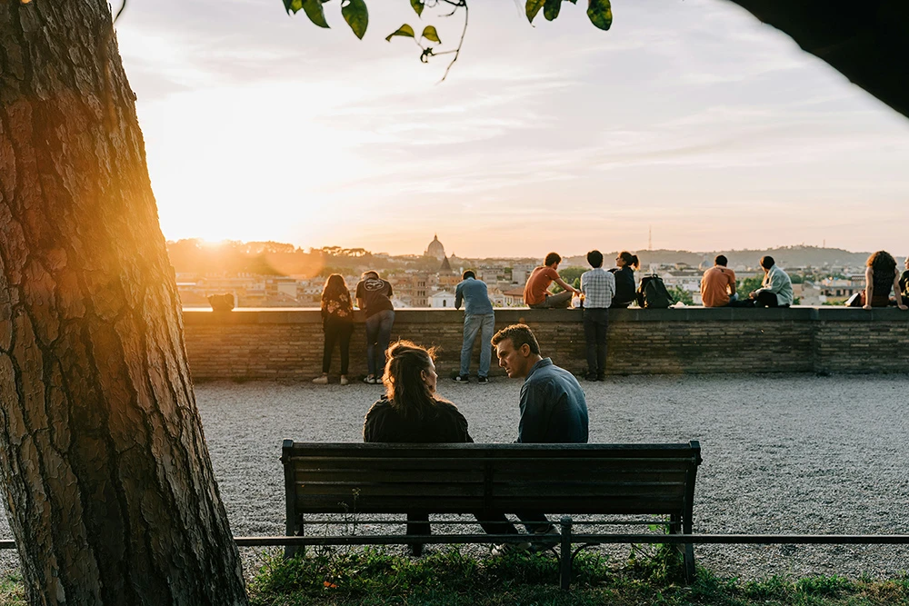 A couple sits on a bench at sunset, talking quietly, while others gather along a stone wall overlooking the city.