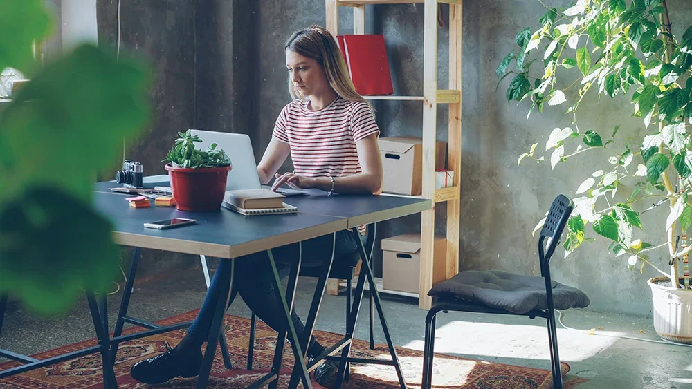 A person sits at a desk working on a laptop in a bright, plant-filled home office.