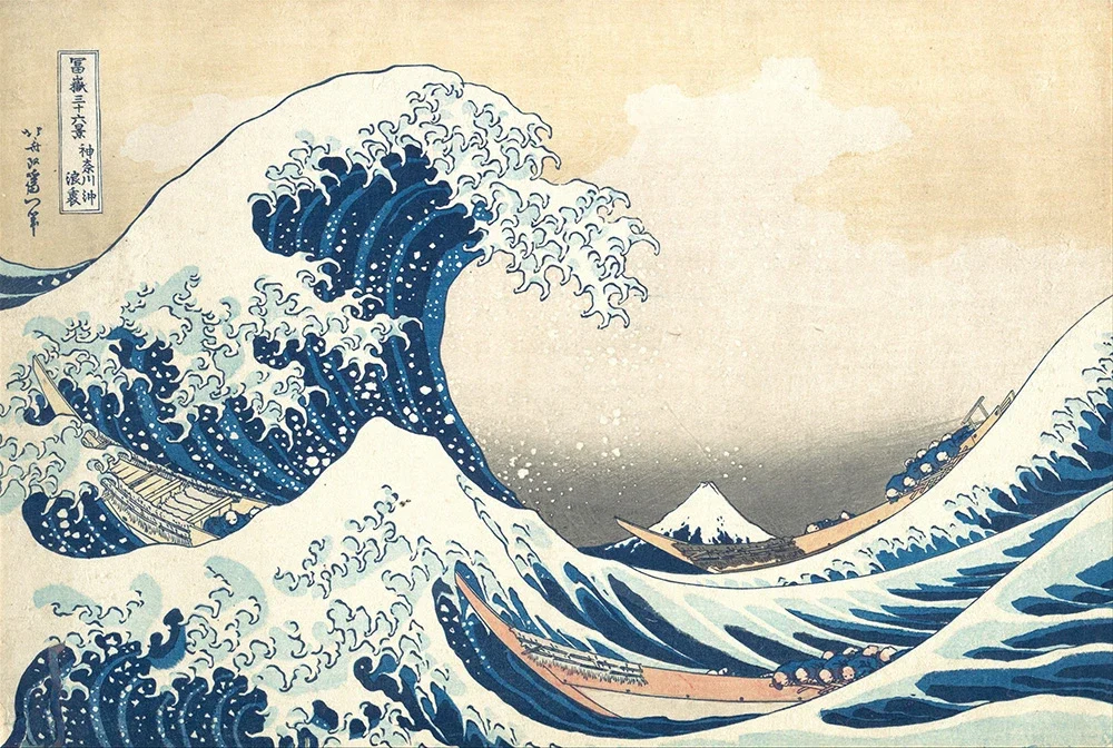 A vibrant Japanese woodblock print showing towering dark blue waves framing a distant Mount Fuji. Three small boats with rowers are tossed about in the turbulent water below.
