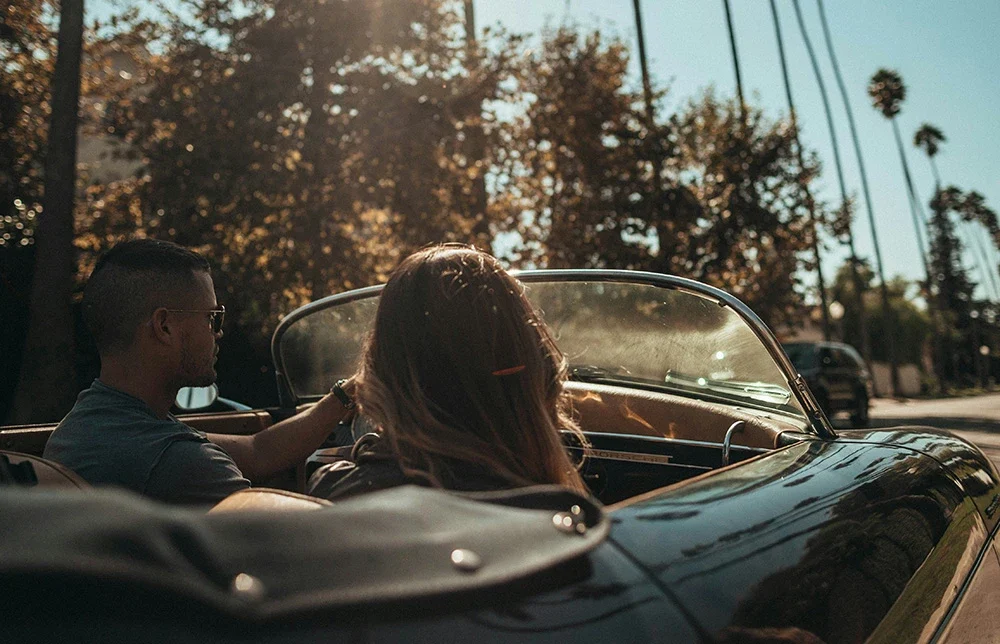 A couple sit in a vintage convertible, driving through a sunlit street lined with palm trees on a warm afternoon.