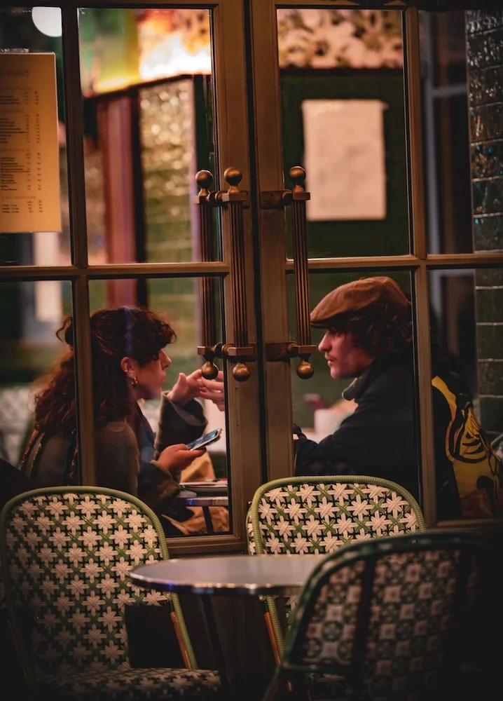 Two people in conversation at a café table, seen through a paned glass door, with woven bistro chairs in the foreground.